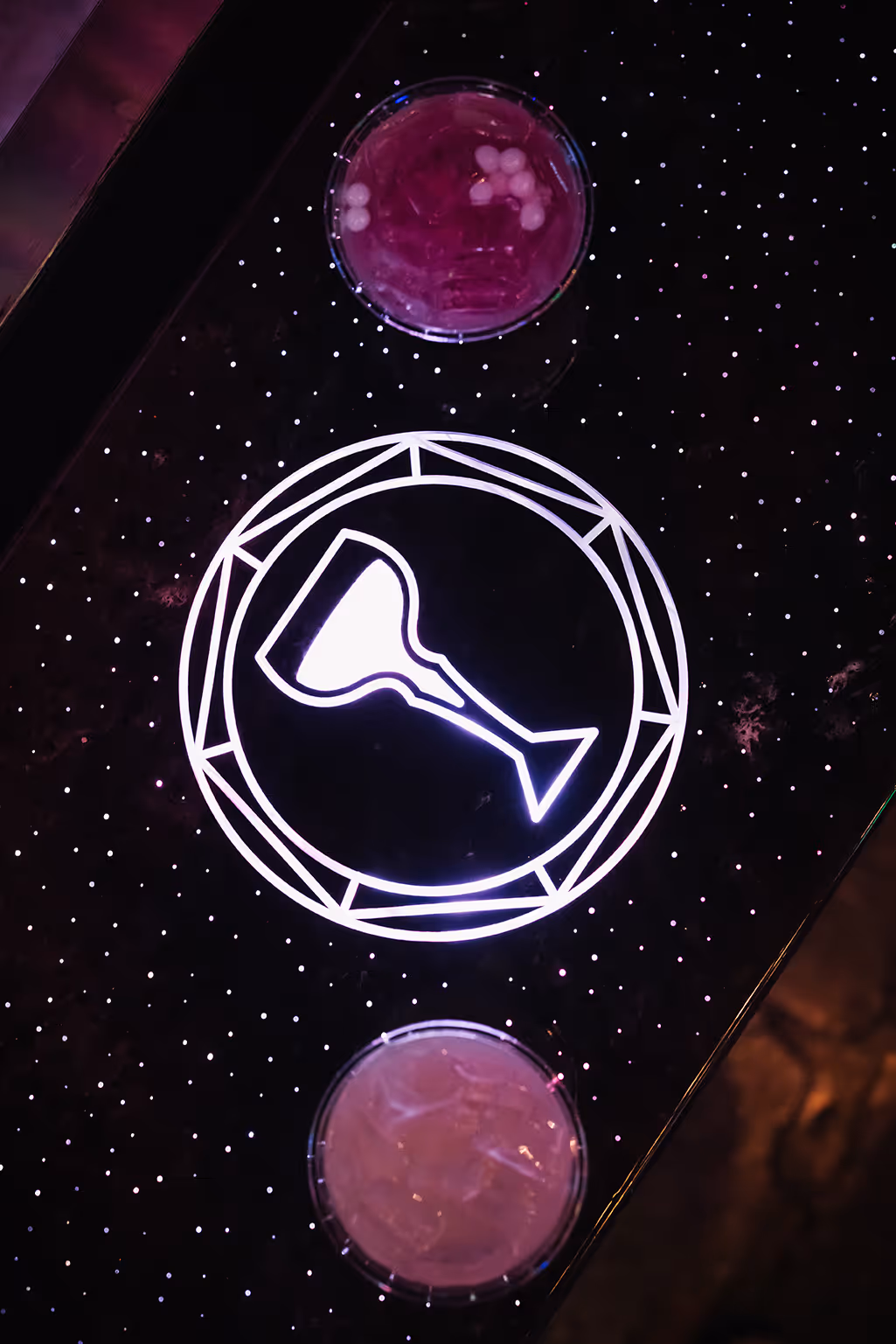 Two purple cocktails on a dark table with a glowing white wine glass icon in the center.