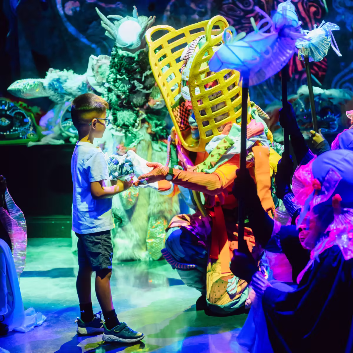 A strange character with a neon yellow head piece extends a hand to a child who is visiting Meow Wolf Houston. 